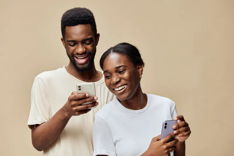 Joyful young African couple engaging with smartphones, showcasing happiness and connection against a warm beige background