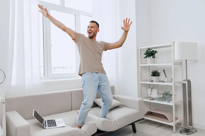 Happy man celebrating work-life balance, embracing joyful moments at home while standing on a couch with a laptop, enjoying life and freedom from stress.