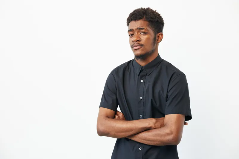 Confident black man with crossed arms, wearing a sleek black shirt against a plain background. Ideal for conveying strength and personality in modern settings.