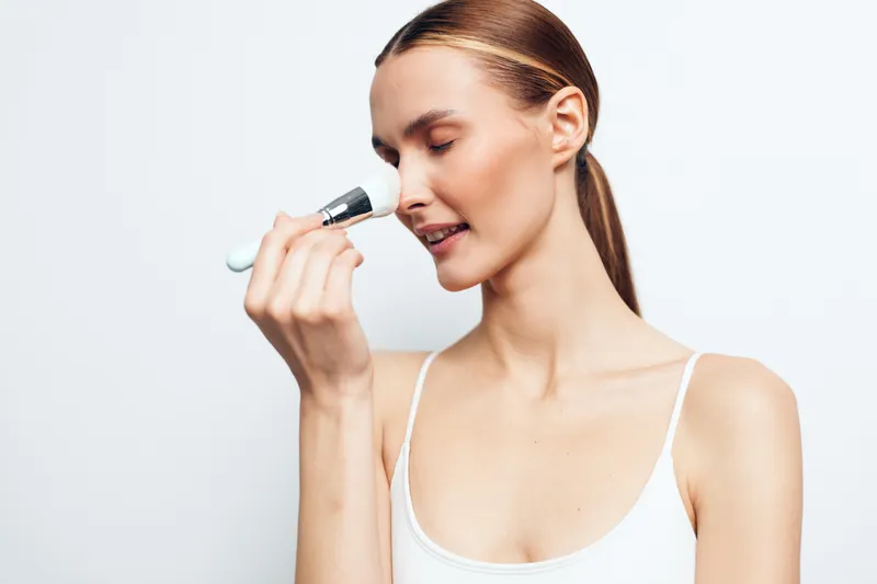Closeup of a woman applying makeup with a brush, soft light, serene expression, fresh complexion, minimal style, white background, beauty routine in clear detail and natural tones