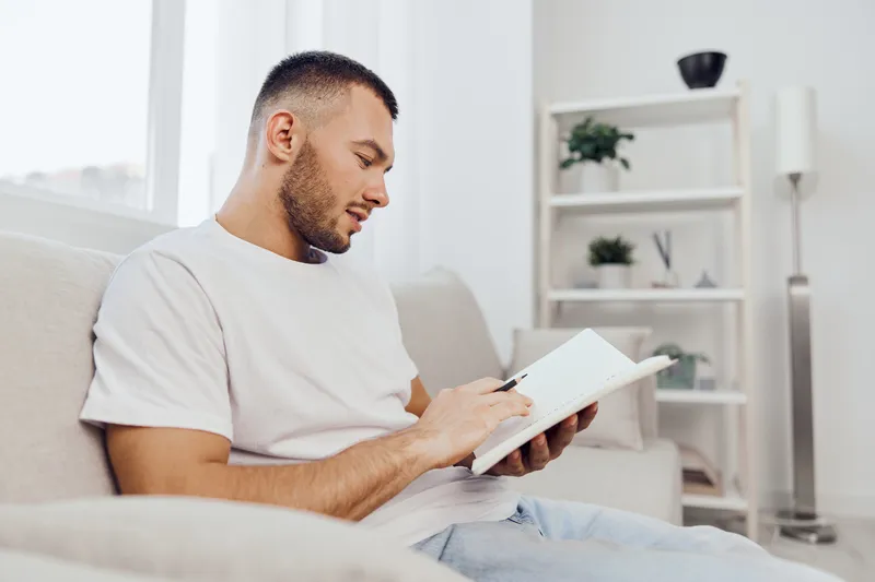 Thoughtful man writing in a notebook while sitting on a cozy sofa, surrounded by modern decor. A perfect blend of creativity and comfort for daily inspiration.