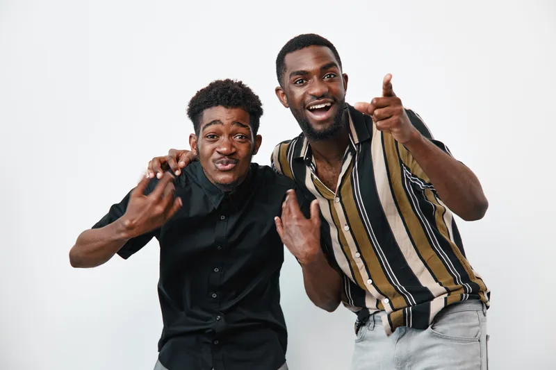 Two young black men with dark skin posing together on white background, smiling and making hand gestures, casual clothing and joyful expressions in studio.