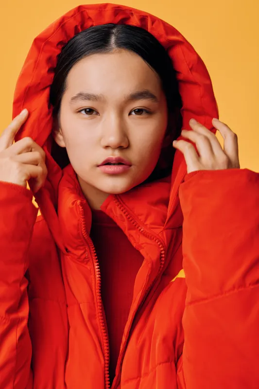 Young woman wearing red hooded jacket, portrait with serious expression on yellow background, fashion style and winter clothing with hands adjusting hood in studio lighting.