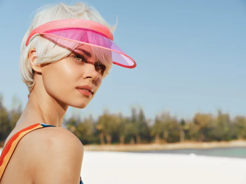 Portrait of a stylish woman at the beach wearing a translucent pink visor, sunlit skin, and bright swimsuit strap, capturing a carefree summer vibe.