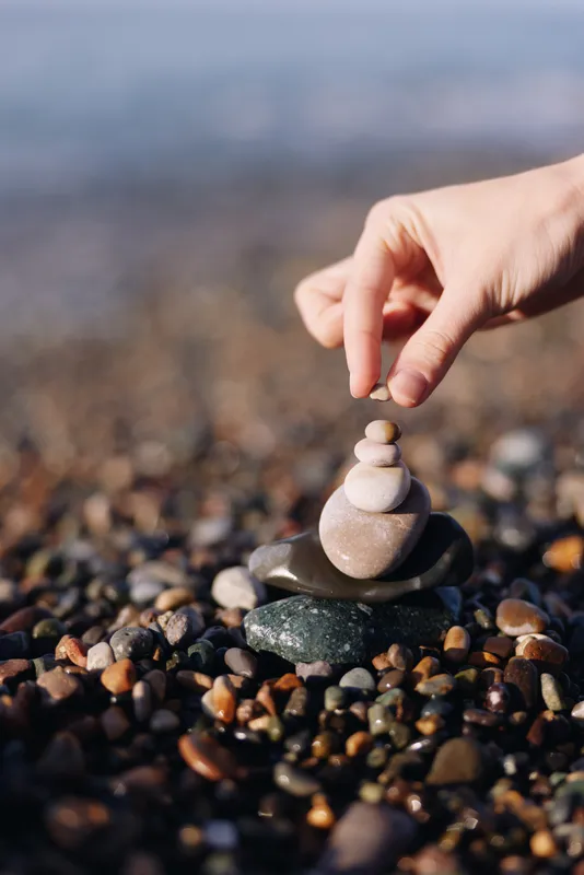 Close up of a hand placing rounded stones into a small cairn on a pebbled beach, capturing calm balance, focus, and nature’s tranquil mood.