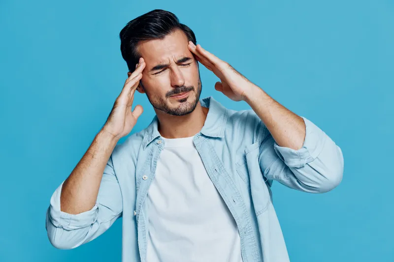 Stressed young caucasian man with headache holds his head, frowning and closing eyes, dressed in casual light blue shirt, standing against solid blue background.