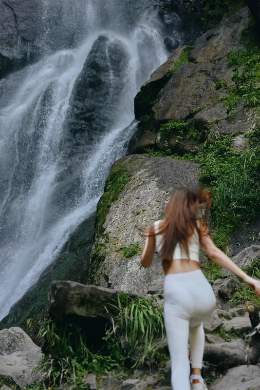 Young woman hiking near a large waterfall in lush green forest, wearing white activewear while exploring rocky terrain and enjoying nature outdoors.
