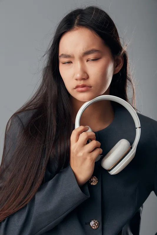 Young woman holding headphones with long dark hair and serious expression on face wearing dark jacket isolated on gray background for music and emotion concept.