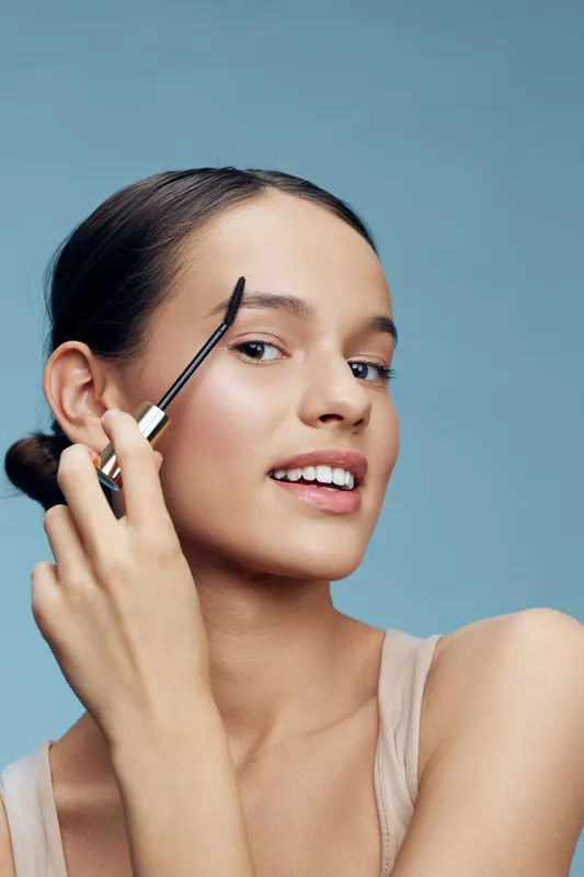 Young woman applying brow makeup against a light blue background, showcasing confidence and beauty with natural skincare and elegant style