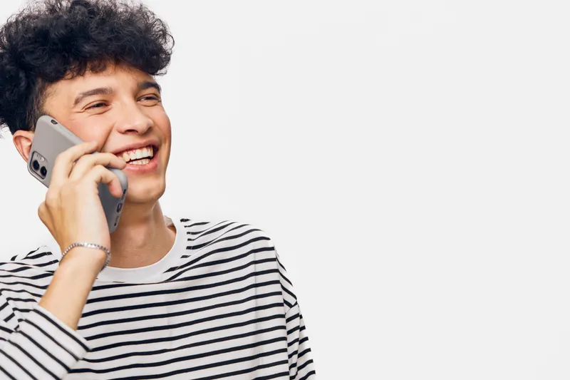 smiling man talking phone, young adult with curly hair wearing striped shirt, cheerful expression, white background, positive communication moment