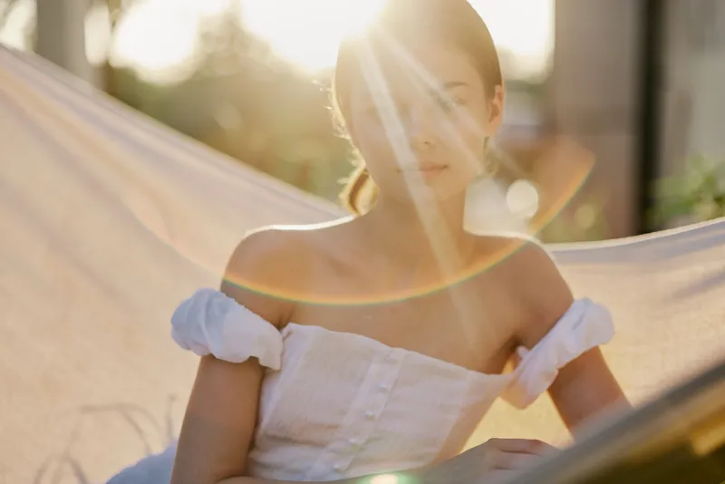 A serene moment of a young woman in a white summer dress, basking in sunlight while relaxing on a hammock, evoking tranquility and summer vibes