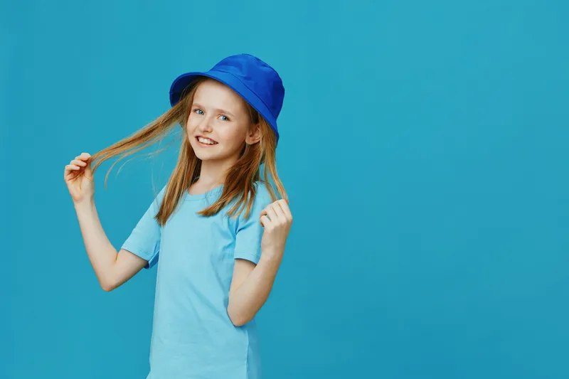 Cheerful girl with long hair in a blue hat, smiling and playing with her hair against a vibrant blue background, perfect for children s fashion themes