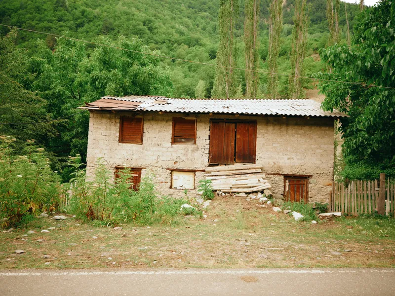 Rustic countryside house with weathered plaster walls, wooden shutters and rough steps, surrounded by greenery and trees, a calm rural dwelling by a quiet country road at noon