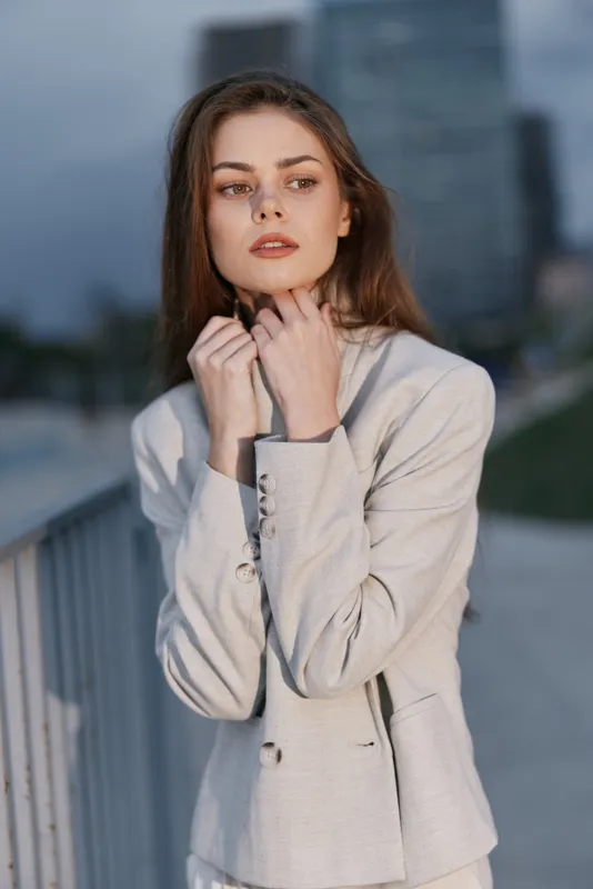 Young woman in light beige jacket posing outdoors at dusk with thoughtful expression, urban background and muted color palette, fashion and psychological concept
