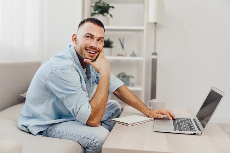 Smiling man working from home, cozy interior, casual attire, modern design, positive vibes, focused on laptop, creativity in action.