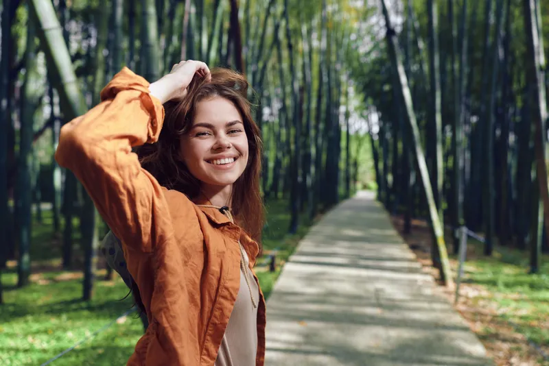 Woman smiling portrait on walkway in bamboo forest, outdoors, happy young adult in orange jacket posing by path, nature background, candid lifestyle and travel mood.
