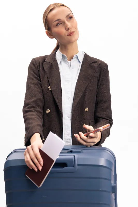 Young woman in brown blazer holds passport and smartphone while standing behind blue suitcase. Concept of travel preparation, business trip, and modern lifestyle with essential documents.