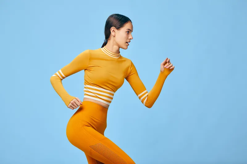 Woman fitness portrait: sporty female in orange seamless activewear captured mid-run pose in a studio. Sport concept highlights toned muscles, focused expression and clean light blue backdrop.