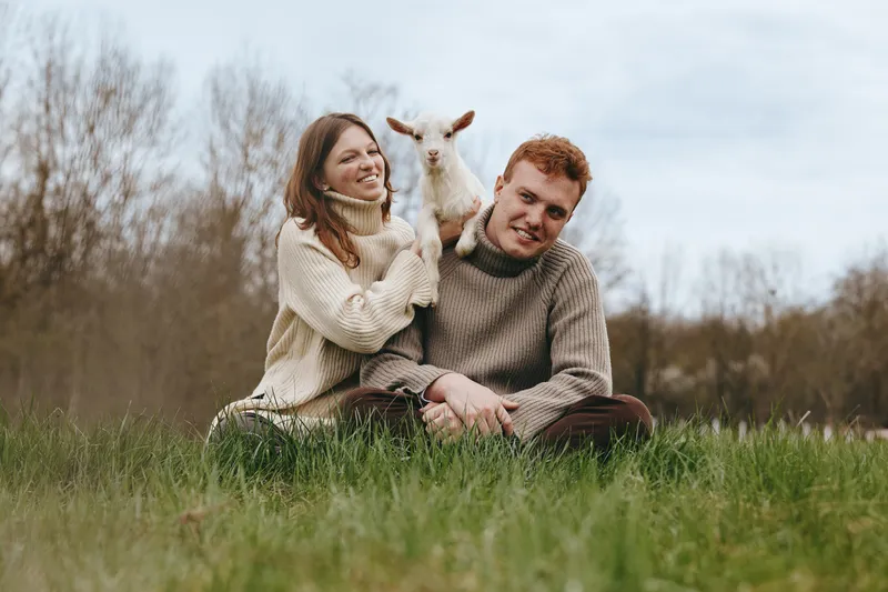 Smiling couple sitting on grass in an open field with a playful goat kid between them, capturing a warm lifestyle moment in nature and rustic charm