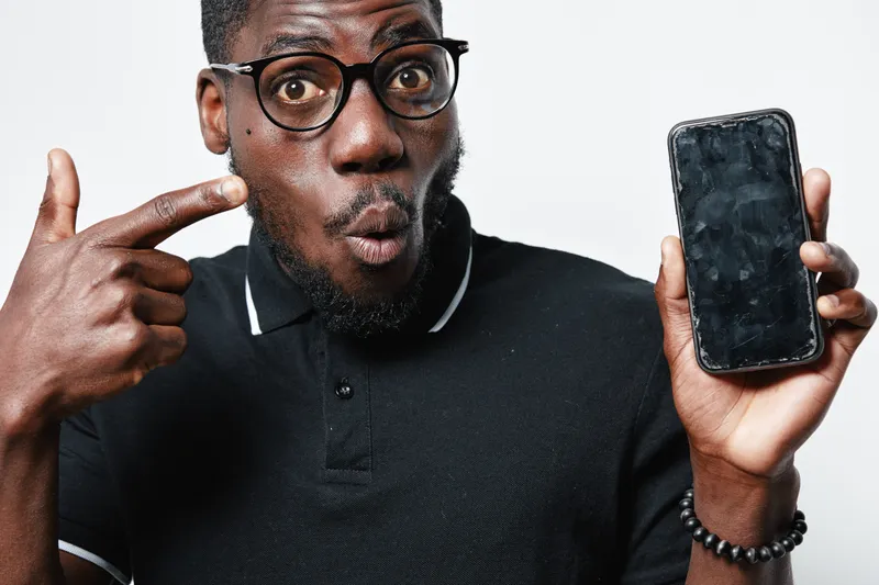 African American man with dark skin and glasses pointing at smartphone with cracked screen, surprised expression on face, wearing black polo shirt.