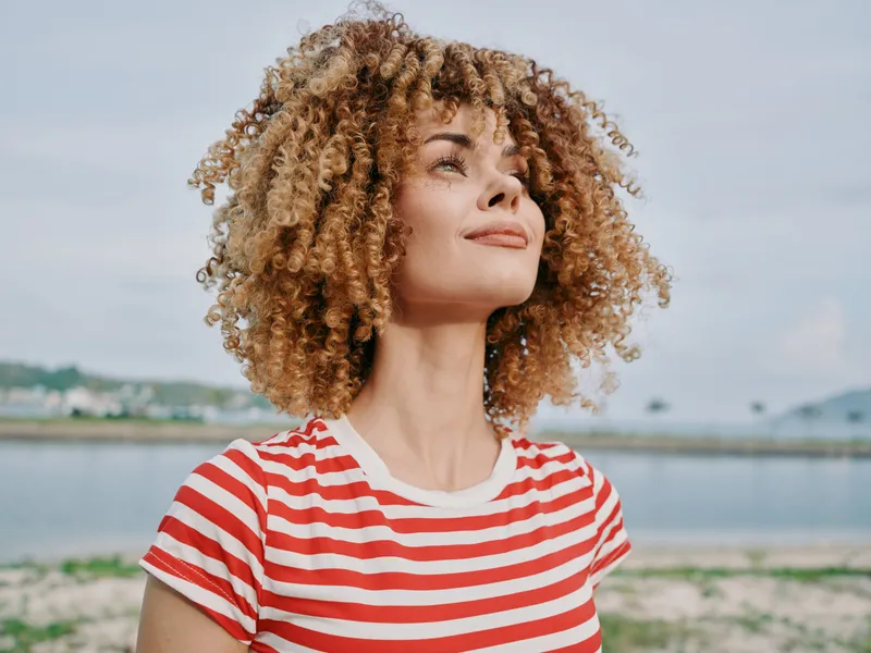 Portrait of a young woman with curly hair, wearing a red and white striped shirt, facing slightly upward near a calm shoreline under a clear sky.