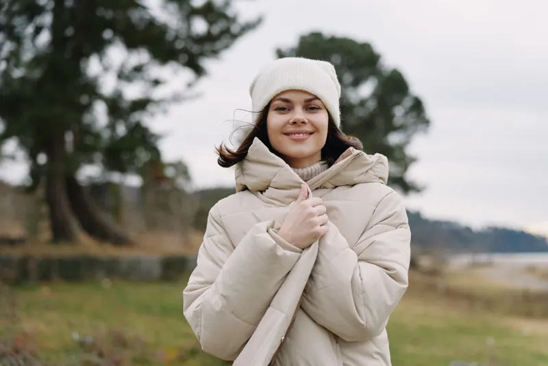 Winter outdoor portrait of a smiling woman in a warm coat, scarf and beanie, beside a shoreline with calm water and trees on a chilly day.