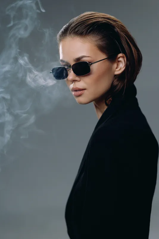 Stylish woman wearing sunglasses with smoky background, expressing cool and confident attitude in dark clothing.