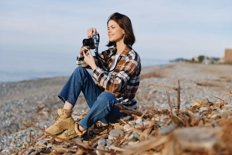 Photographer, woman with camera on a pebble beach, casual plaid shirt, jeans, hiking boots, capturing coastal scenery under clear sky, with wind in hair and calm ocean in the distance.