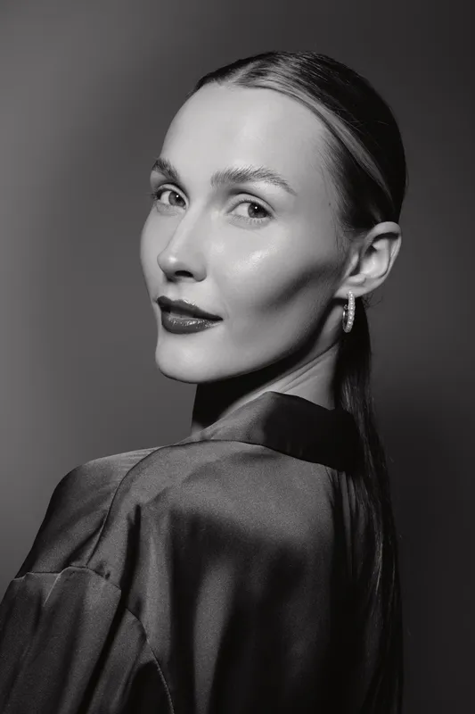 Portrait of a slender Caucasian woman with fair skin and slicked back hair wearing a silky blouse. Studio lighting highlights her smooth complexion and elegant pose.