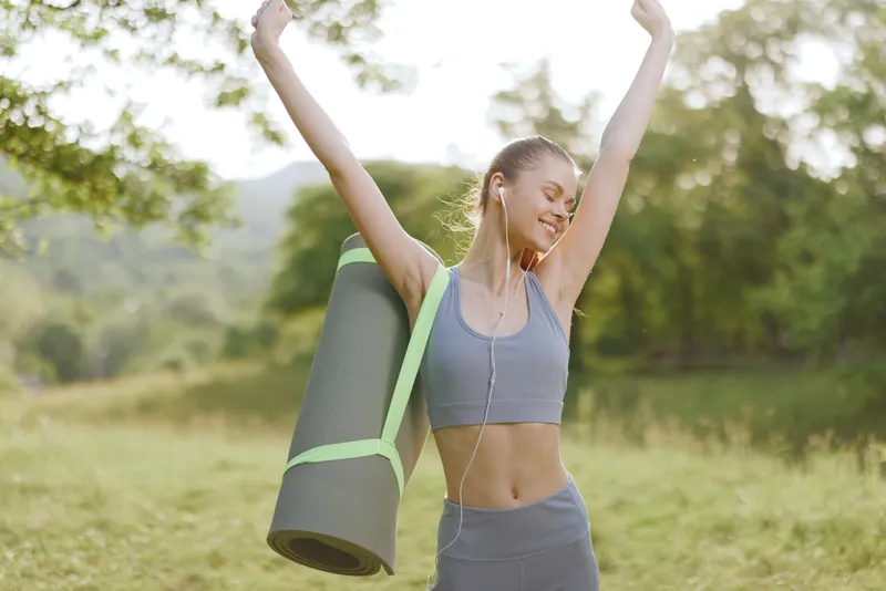 Happy young woman stretching with yoga mat outdoors, embodying the concept of healthy lifestyle and fitness in a vibrant nature setting