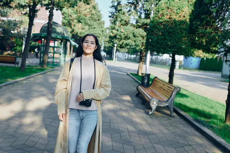 Park, woman, coat, casual, street, fashion, sunlight, trees, bench, stroll in a stylish outdoor moment with a long beige coat and jeans in a bright daytime setting