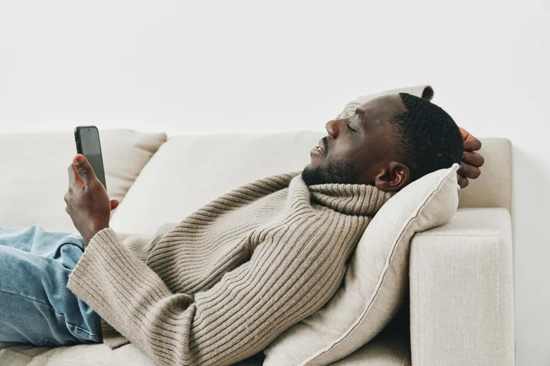 Relaxed young Black man in a cream sweater using a smartphone while lying on a modern beige sofa, showcasing comfort and leisure