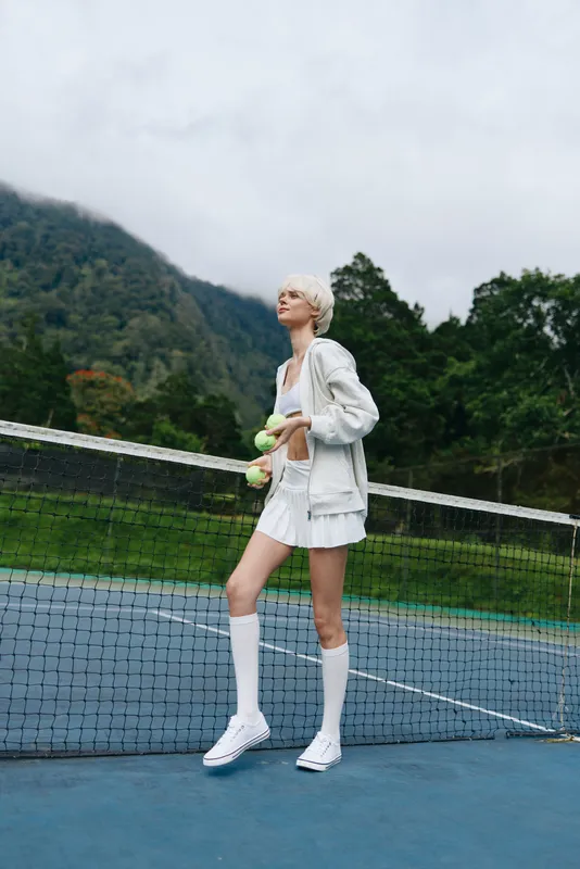 Female tennis player in sporty white outfit holding a tennis ball on a court, showcasing athleticism and confidence against a natural background