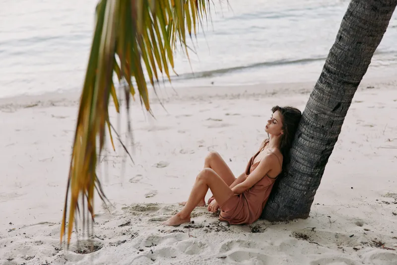 A woman sits relaxed against a palm tree on a sandy beach, wearing a pink dress, bathed in warm light with a calm sea in the background, capturing a tranquil tropical moment.
