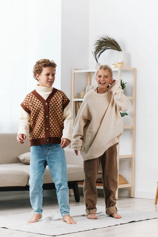 Two children wearing oversized knit sweaters dance and laugh in a bright living room, soft natural light, cozy textures, and a cheerful, carefree moment.