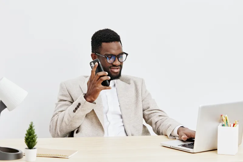 Professional young African American man in smart casual attire working at modern office desk, talking on mobile phone, using laptop, smiling, confident, with minimal decor, indoor workspace, natural l