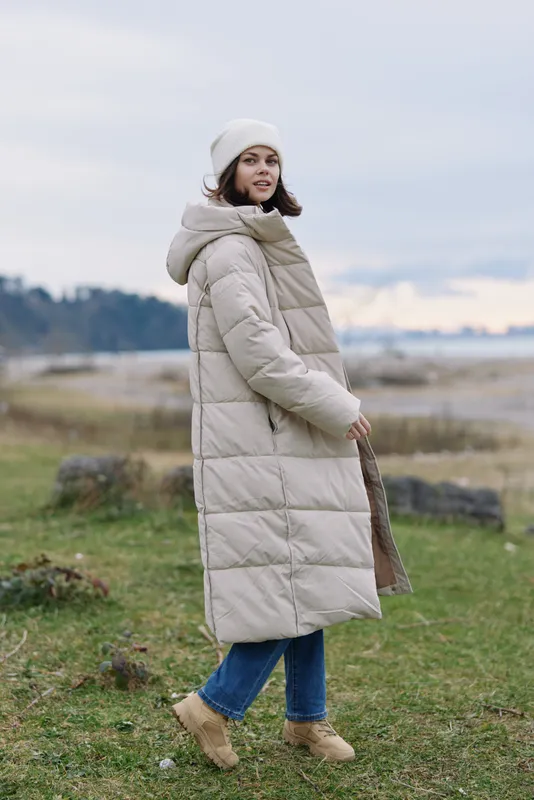 Woman in a long beige puffer coat, beanie, and jeans walking along a windy coastal field with soft clouds, distant shore, and calm atmosphere.