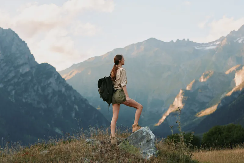Woman hiker with backpack standing on a rock in alpine mountains, exploring nature, casual outdoor clothing, peaceful landscape, bright sky, travel lifestyle, solitary adventure