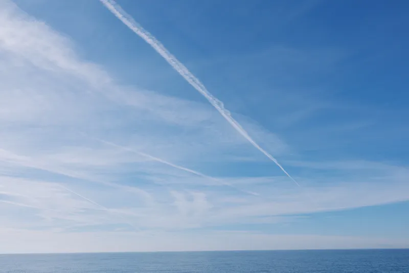 Clear blue sky over calm sea with wispy clouds and a high contrail crossing the horizon, creating a peaceful seascape perfect for travel or nature themes