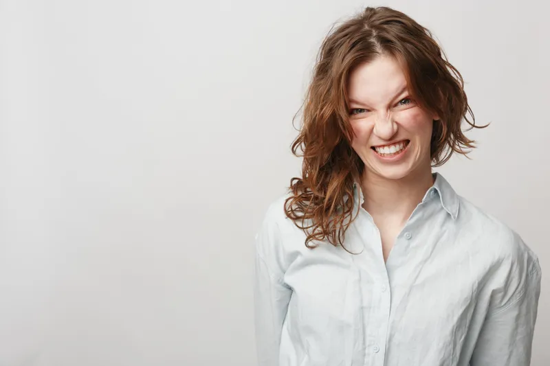 Young woman with curly hair makes playful grimace wearing white shirt on light background. Concept of fun, emotions, youth, and casual lifestyle portrait with natural expression.
