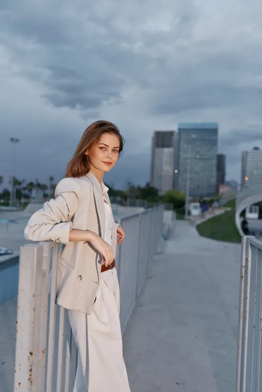 Young woman in beige business suit standing outdoors with skyscrapers background, confident professional woman in urban setting at dusk, business fashion concept