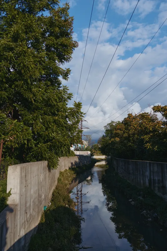 Concrete canal, drainage channel, power lines, birds on wire, trees, reflection, sky, urban nature — Empty suburban drainage with photographer shadow, late afternoon light and moody atmosphere.