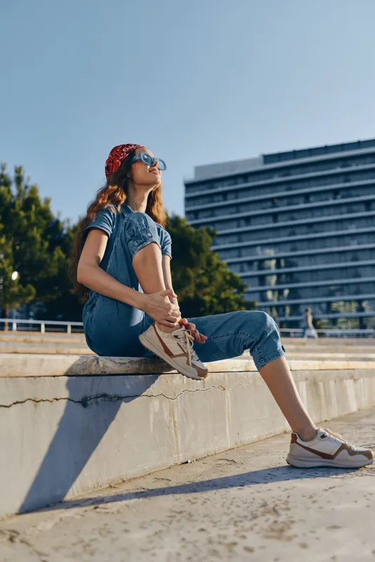 Lifestyle woman in modern boho-western denim street style wearing a red bandana and sunglasses, enjoying sunny urban outdoors with relaxed posture and film color effect.