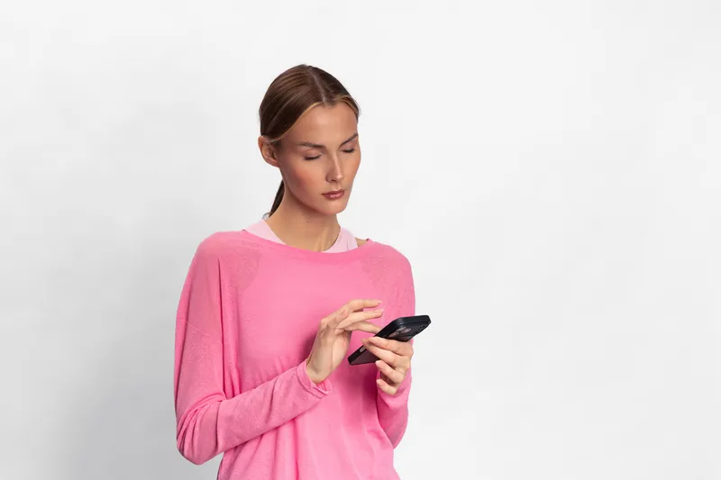 Young woman in pink sweater uses smartphone with focused expression, standing against plain white background. Concept of communication, technology, and modern lifestyle.
