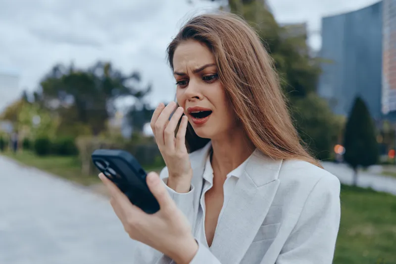 Professional woman expressing concern while using smartphone in urban setting, dressed in formal attire with neutral color palette, showcasing emotional engagement.