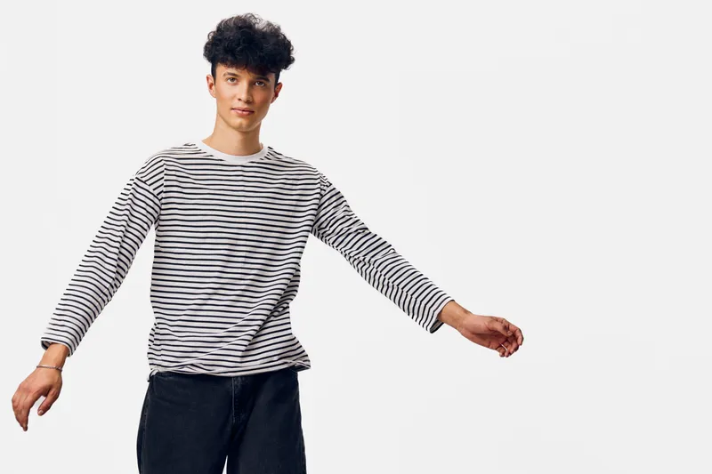 young man wearing striped long sleeve shirt and black pants posing with raised arms against white background in casual style