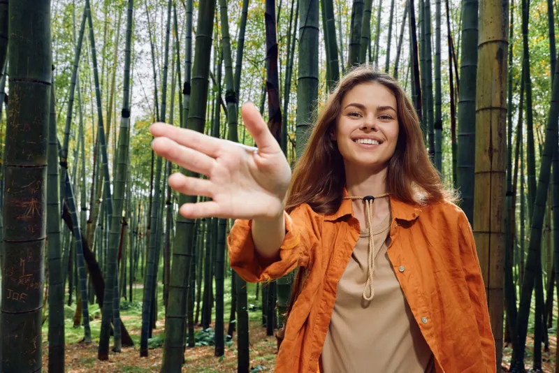 Woman smiling in bamboo forest reaching hand forward in friendly gesture, portrait of young female with orange jacket among tall bamboo trees, nature walk and connection.