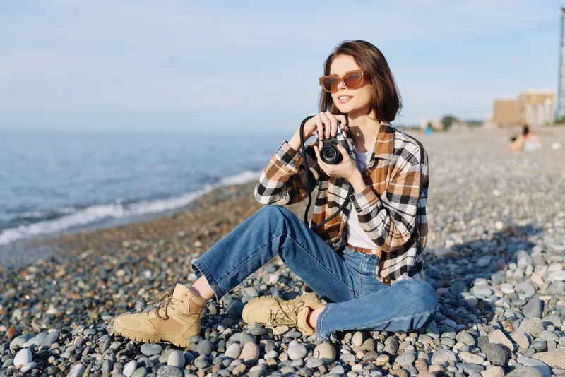 Photographer woman on rocky beach wearing plaid shirt, jeans and boots, holding camera to capture sea scenery, sunglasses on, bright daylight, relaxed outdoor lifestyle portrait with ocean horizon.
