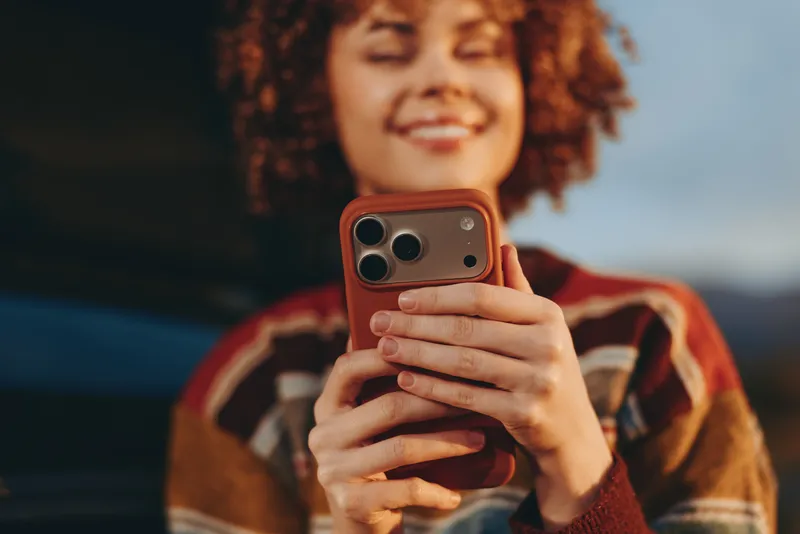 Woman lifestyle portrait indoors, curly hair framing her face as she holds a smartphone for a selfie, relaxed smile, casual sweater and visible hands, soft blurred couch background, warm light.