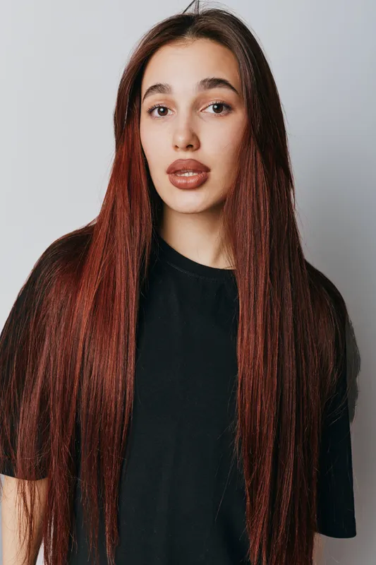 Woman with long straight hair and natural makeup wearing black t shirt standing against white background looking at camera with neutral expression for portrait or beauty concept.